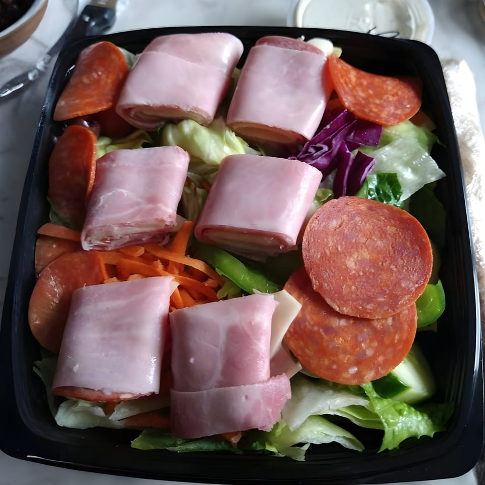 A fresh salad with rolled ham and cheese, pepperoni, lettuce, and other vegetables in a black container.